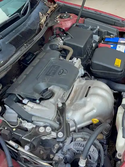 Engine bay of a red Toyota Camry before cleaning, with dust and grime across the engine cover and surrounding panels