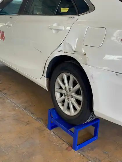 Rear quarter panel of a white Camry raised on stands, dented and creased above the rear wheel arch