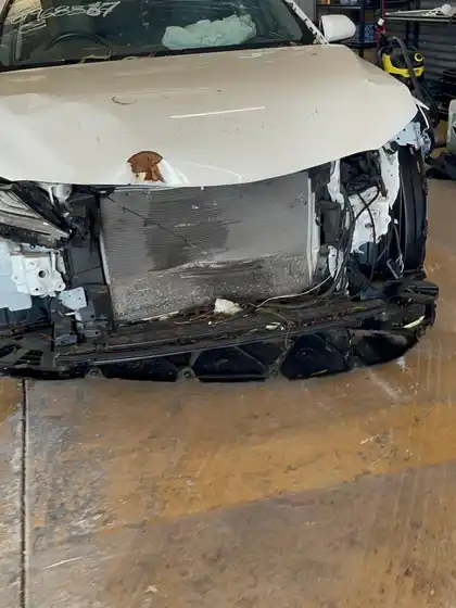 Front of a white Toyota Camry after a collision, with the bonnet buckled, the front bumper torn away and the radiator support exposed