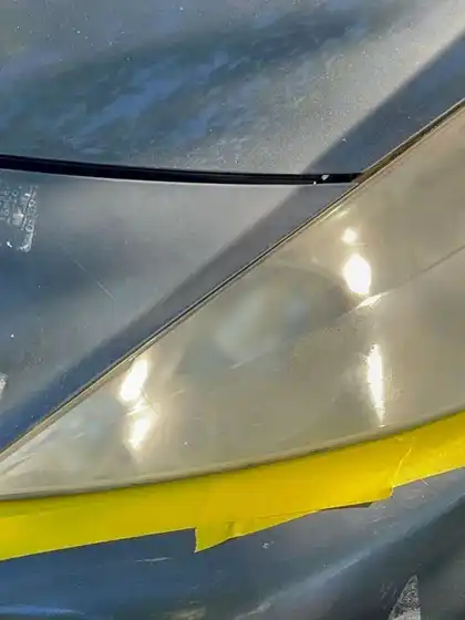 Close view of a car headlight before restoration, the lens clouded and yellowed with the surrounding paint masked off